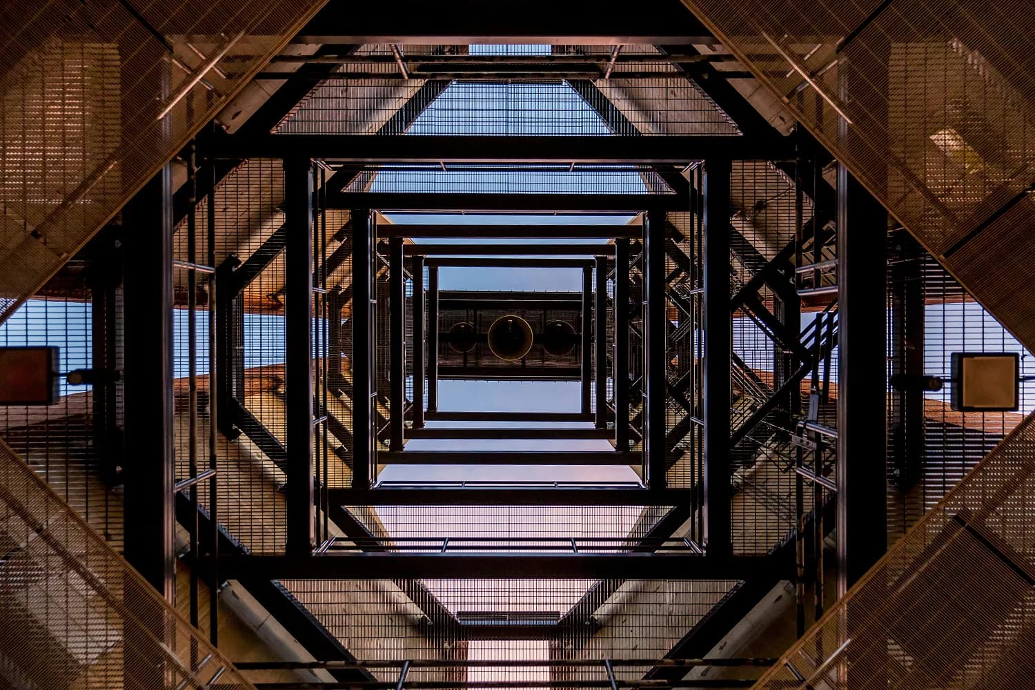 Looking up through a square metal atrium with repeating frames and a central opening to the sky