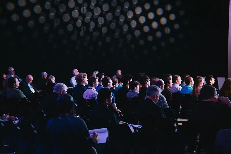 Audience seated in a dark conference hall, facing the stage