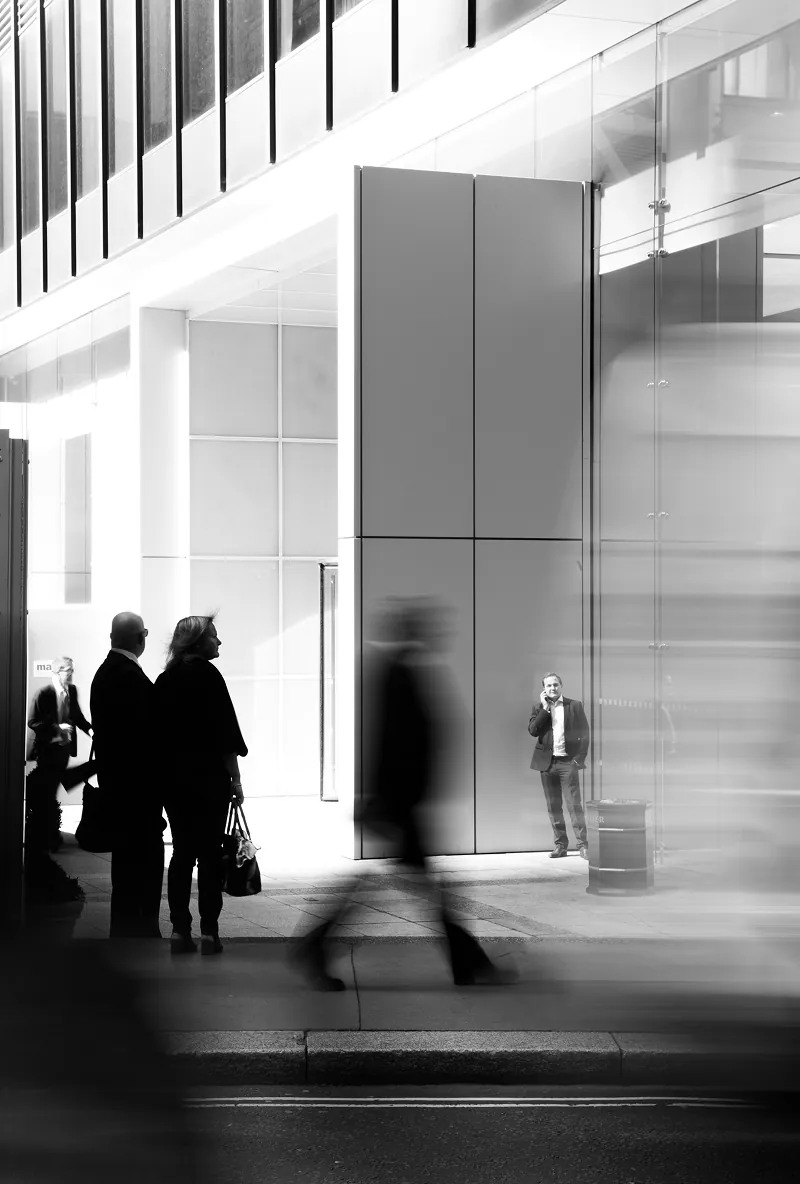 People standing and walking outside a modern glass building, with one man on a phone